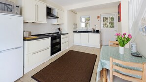 A compact kitchen with white cabinets, a black stove, and a microwave. There's a small dining area with a table and chairs, and a window with a view of the outside. - Cosy cottage in sleepy village yet only 5 minute drive to Truro city centre.  (Malpas)