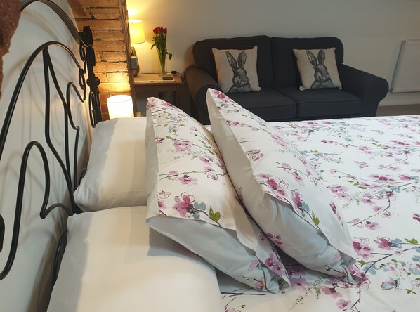 Una cama con ropa de cama floral, un sofá oscuro con almohadones con motivos de conejos y una mesita de noche con una lámpara y un jarrón de rosas rojas.