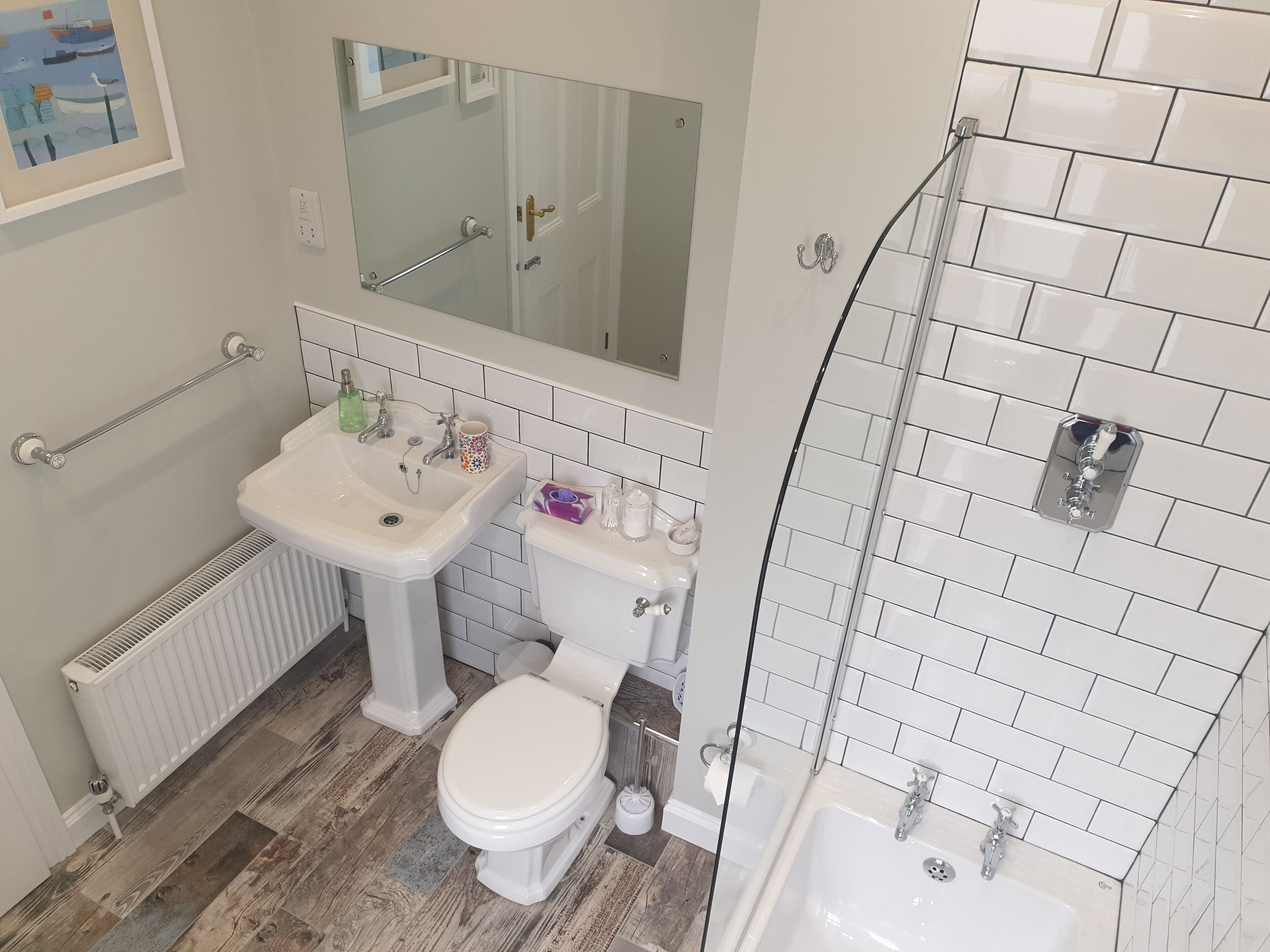 Un baño con lavamanos blanco, inodoro y bañera, suelo de madera y un espejo sobre el lavamanos.