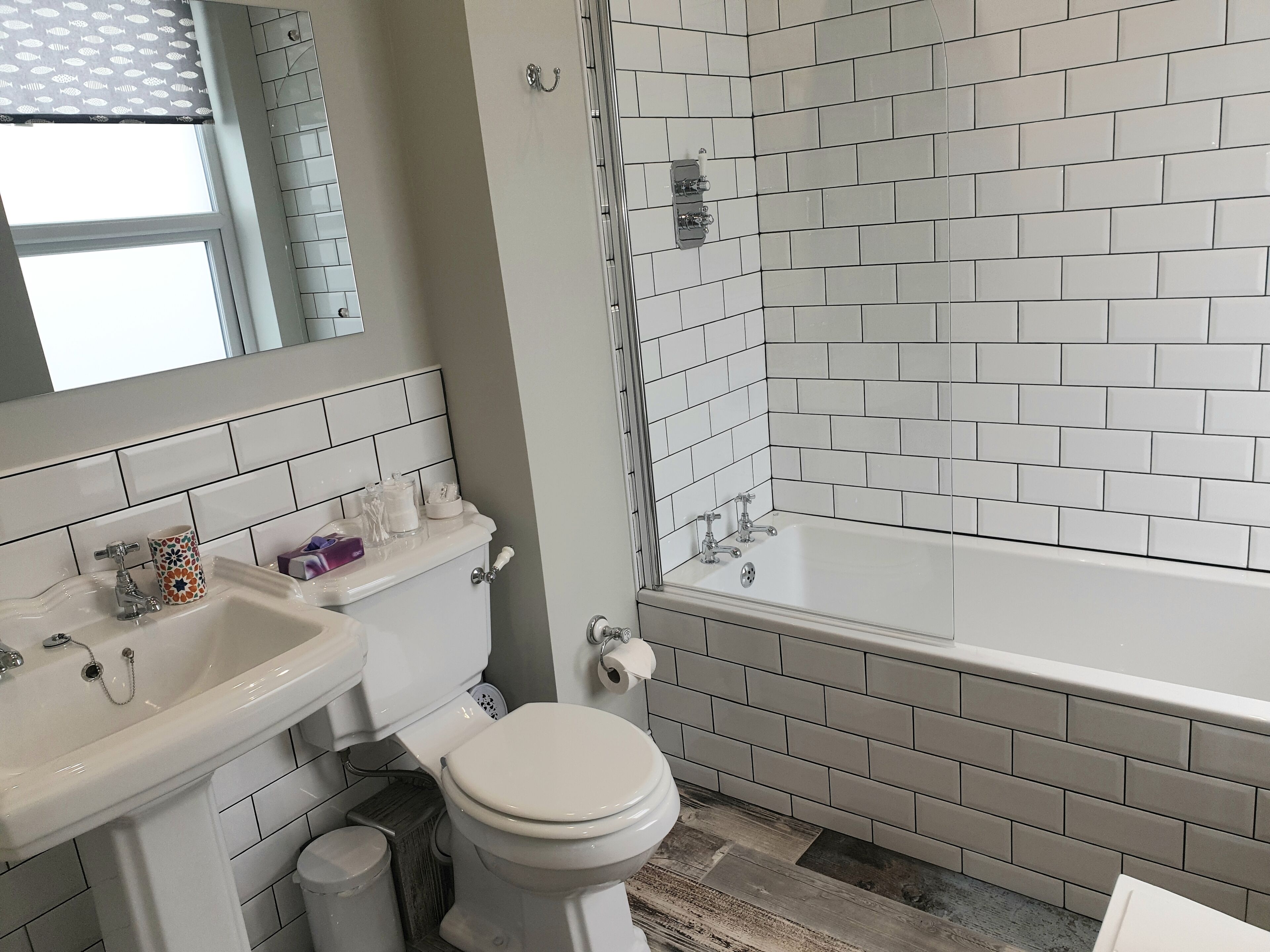 Une salle de bain avec des carreaux de céramique blancs rectangulaires, des toilettes blanches, un lavabo avec un miroir et une baignoire.