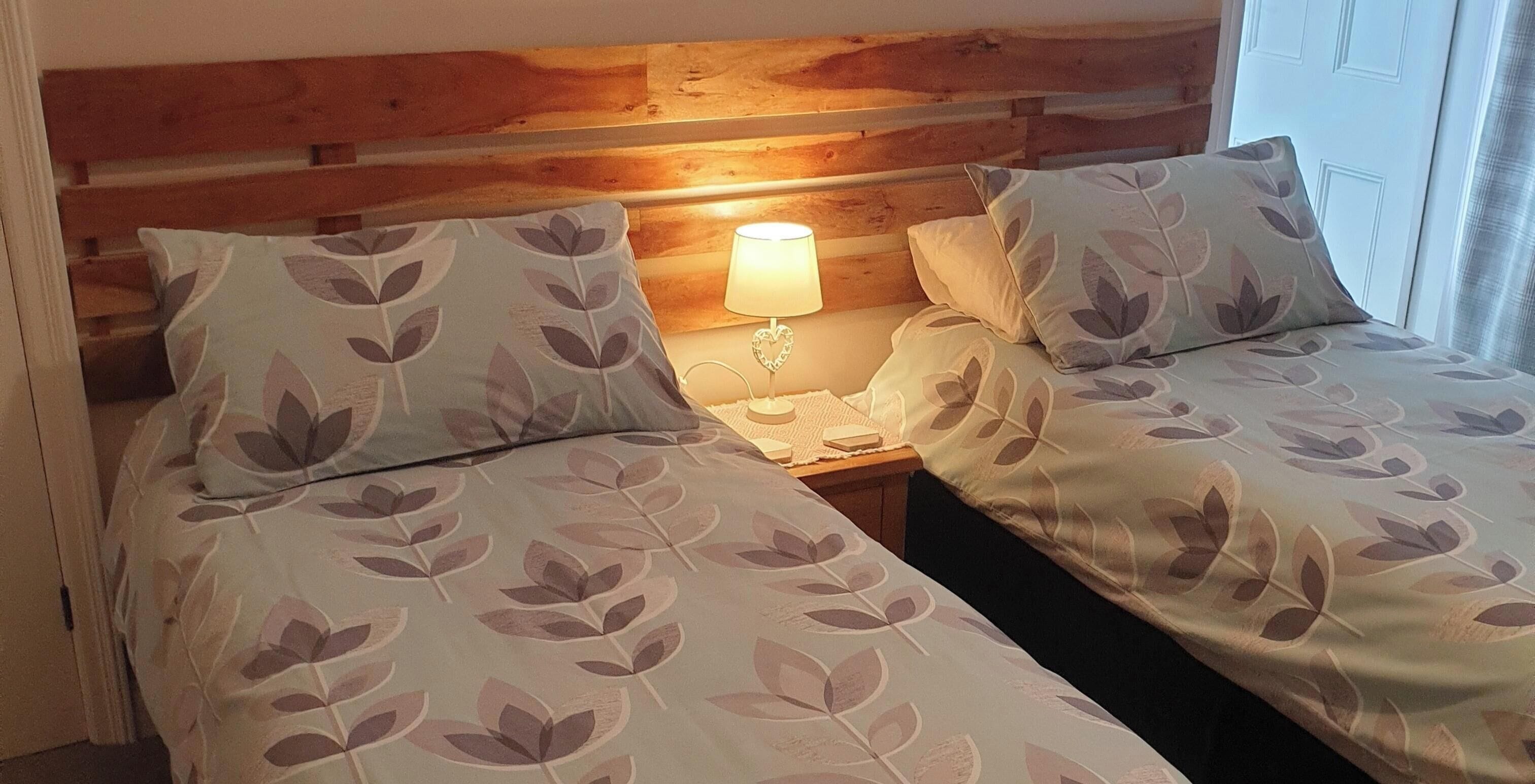 A room with two beds, each with leaf-patterned bedding, a wooden headboard, and a bedside table with a lamp.