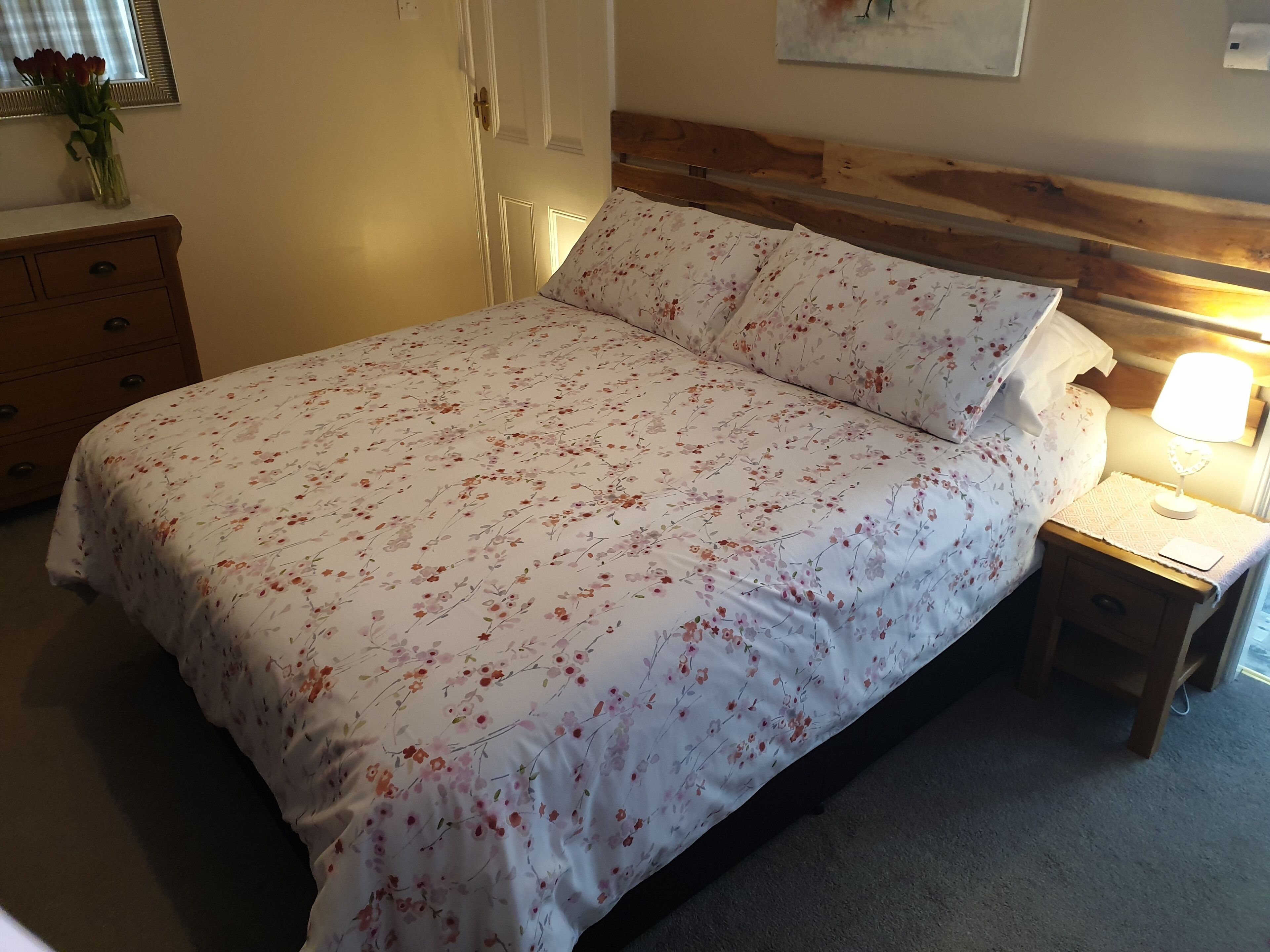 A neatly made bed with floral bedding, a wooden headboard, a bedside lamp, and a nightstand with a small table.