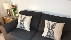 Un sofá gris oscuro con dos cojines estampados con conejos, una mesita de noche con una lámpara y un jarrón de flores, y una pared lisa de fondo.