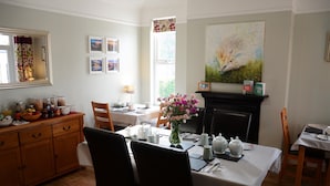 Un comedor con una mesa preparada para dos personas, un jarrón de flores y una chimenea. Hay marcos de cuadros en la pared y una ventana con cortinas.