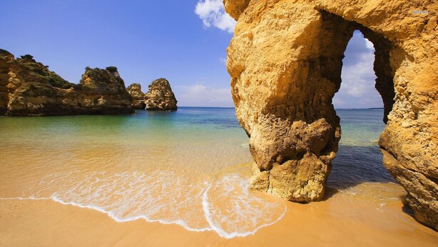 Une plage à l’eau turquoise et limpide, un rivage sablonneux et une grande formation rocheuse surmontée d’une arche.