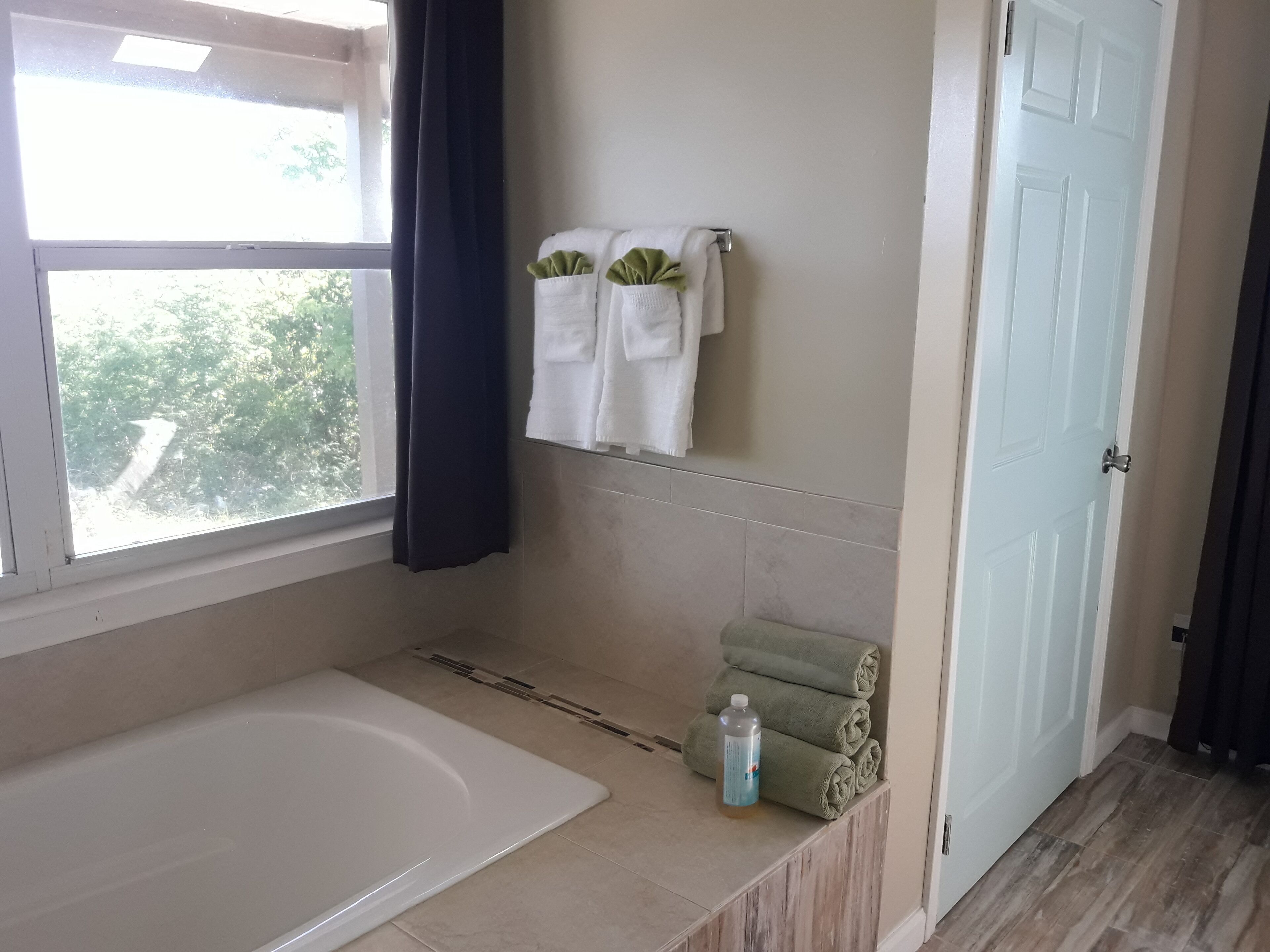 Un baño con bañera, una ventana con cortinas, un perchero con toallas y una puerta.