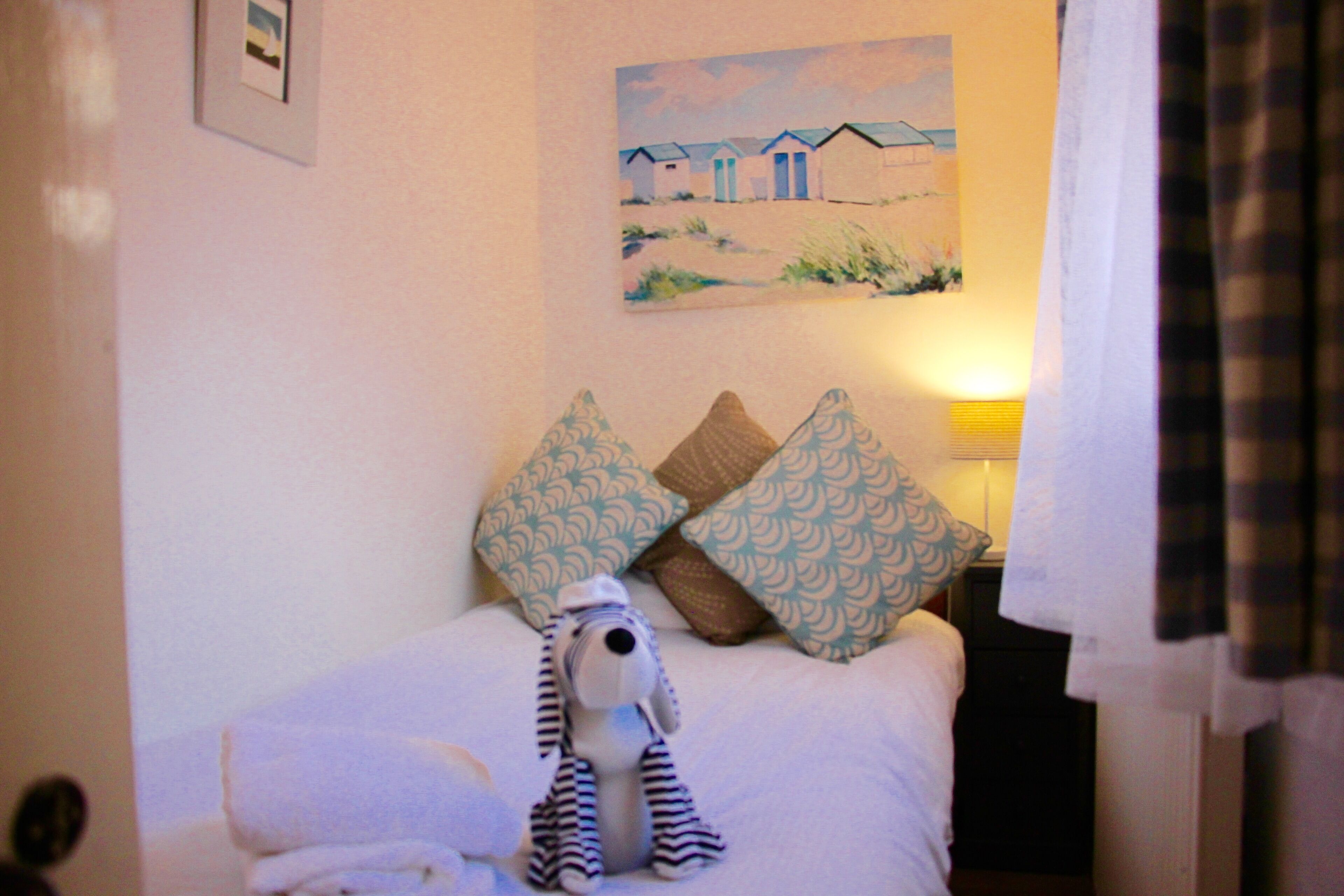A neatly made bed with patterned pillows, a small stuffed toy, and a painting of beach houses on the wall.