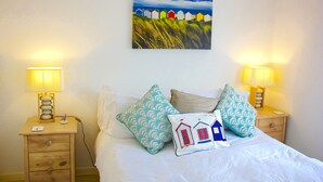 Una cama bien hecha con ropa blanca, dos mesitas de noche con lámparas y un cuadro colgado en la pared de cabañas de playa coloridas.
