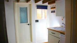 Un pasillo estrecho que conduce a una puerta blanca con una toalla azul a cuadros colgada. Se ve una ventana con una persiana azul, y un pequeño pizarrón con escritura está sujeto a la pared.