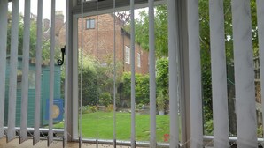 Blick durch ein Fenster mit Metallgitter, ein Ziegelgebäude und ein Garten mit Blumen und Gras.