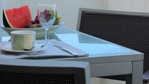 Une table avec un saladier de fruits, un verre d’eau et une tasse, placée sur un balcon d’où l’on peut admirer les bâtiments.