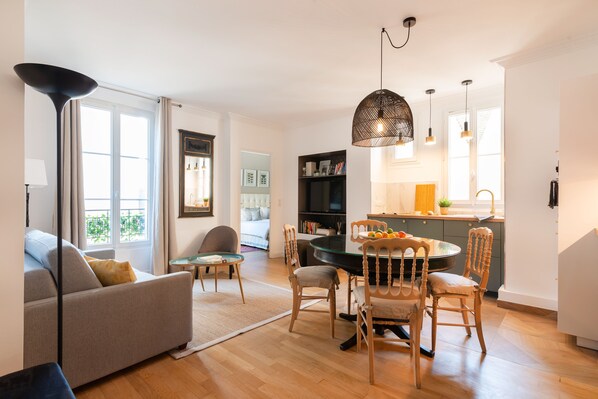 Een moderne woonkamer met een bank, eettafel en stoelen, een keuken met een eettafel, en een groot raam dat zorgt voor daglicht.