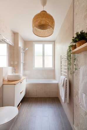 Een moderne badkamer met een vrijstaand bad, een grote spiegel, een witte wastafel en een hangende ratanlamp.