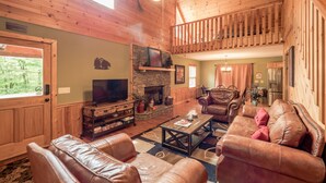 A spacious living room with a fireplace, leather sofas, a TV, and a balcony with wooden railings.
