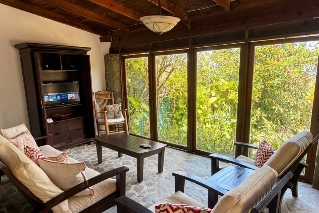 Una habitación con suelo de piedra, televisión, sofá, sillón y ventanas amplias con vistas a zonas verdes.