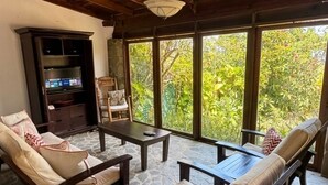 A room with a stone floor, a television, a sofa, a chair, and large windows with a view of greenery.