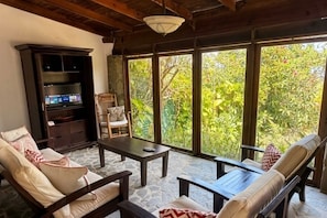 A room with a stone floor, a television, a sofa, a chair, and large windows with a view of greenery.