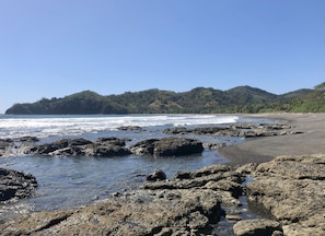 Una costa rocosa con olas rompiendo sobre la playa, una zona arenosa y una colina boscosa al fondo.