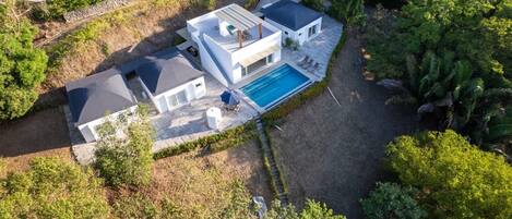 Uma casa moderna de dois andares com piscina, rodeada por vegetação densa.