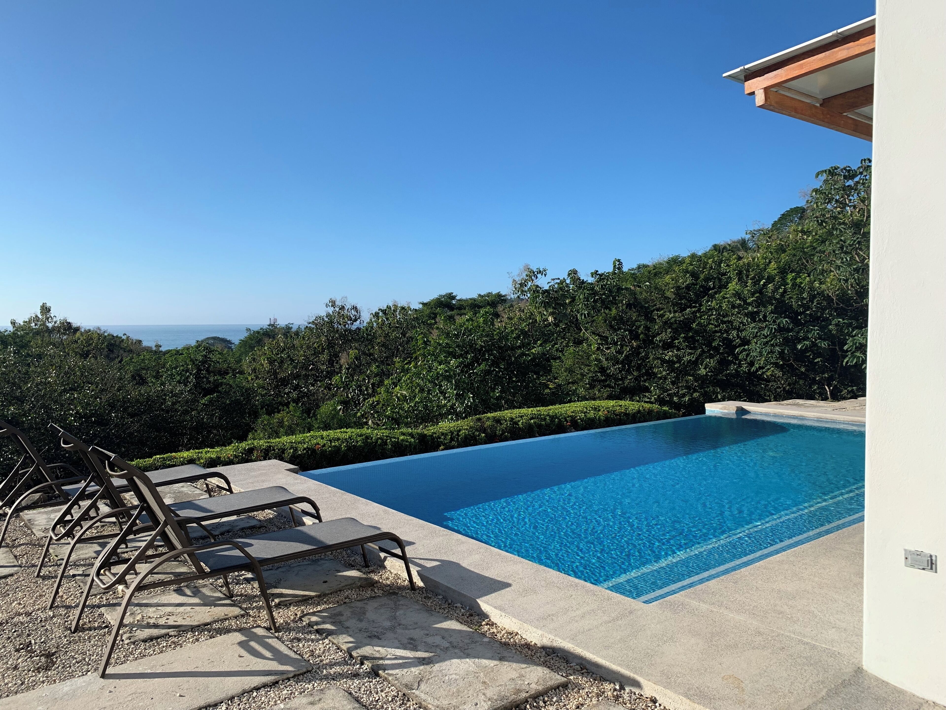Uma piscina azul límpida com espreguiçadeiras, rodeada por vegetação e vista para o mar.