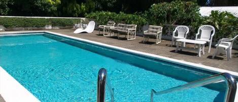 Una piscina rectangular con agua azul cristalina, rodeada por una terraza empavedada. Hay varias sillas de descanso blancas y algunas mesas con sillas en la terraza.