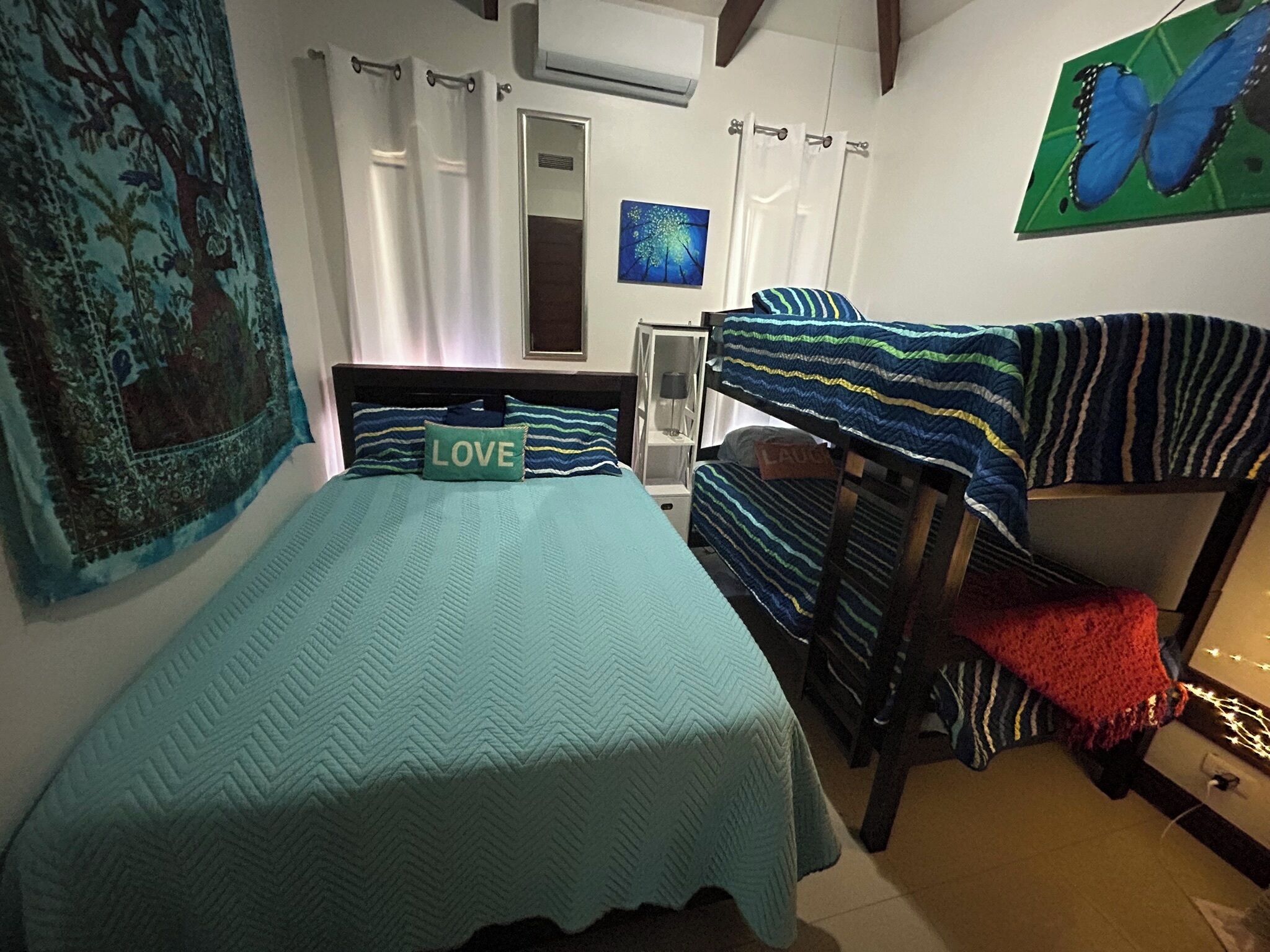 Une chambre à coucher avec un lit, des lits superposés, un climatiseur fixé au mur, une fenêtre avec des rideaux et un tableau.