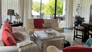 A spacious living room with a ceiling fan, large windows with curtains, a sofa with red cushions, a coffee table, and a dining area with a table and chairs.