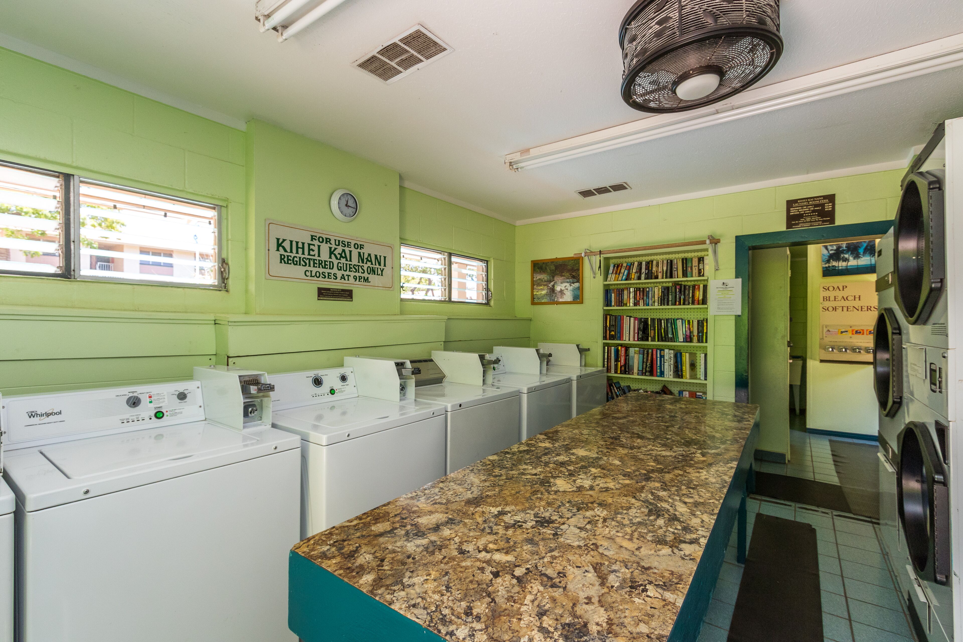 A laundry room with multiple Whirlpool washers and dryers, a green wall with a sign, and a bookshelf with books.