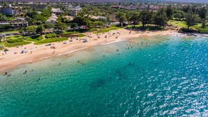 Vista aérea de una playa con aguas turquesas cristalinas, una orilla arenosa y colinas y edificios al fondo.