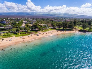 Vista aérea de una playa con aguas turquesas cristalinas, una orilla arenosa y colinas y edificios al fondo.