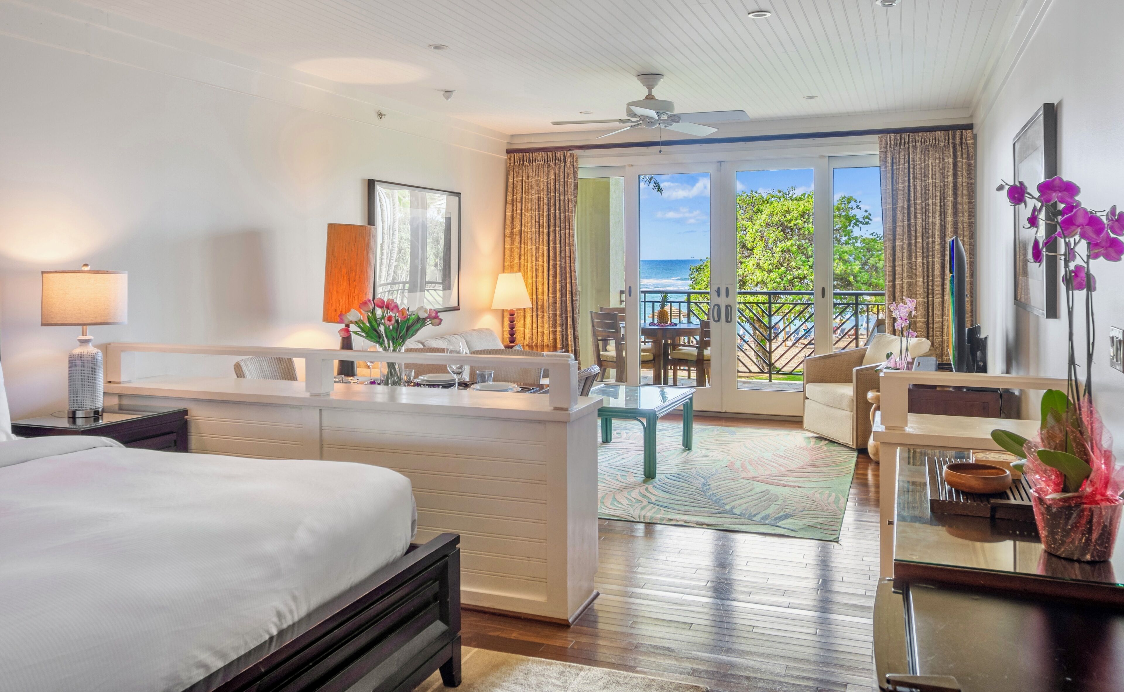 A hotel room with a bed, a desk, a chair, a dining table, and a balcony with a view of the ocean.