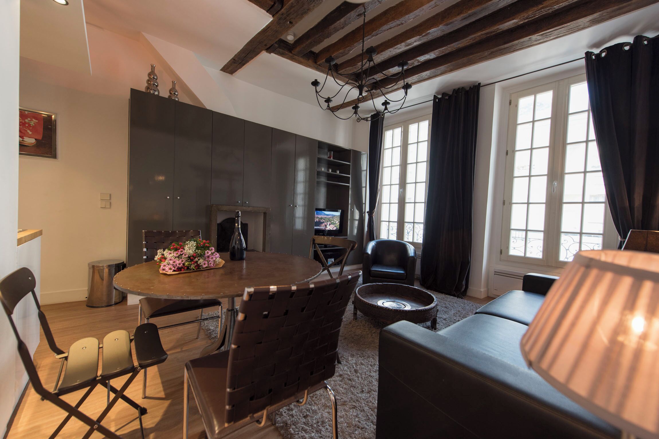 A modern living room with a dining area, featuring a wooden ceiling, a fireplace, a television, and a large window with curtains.