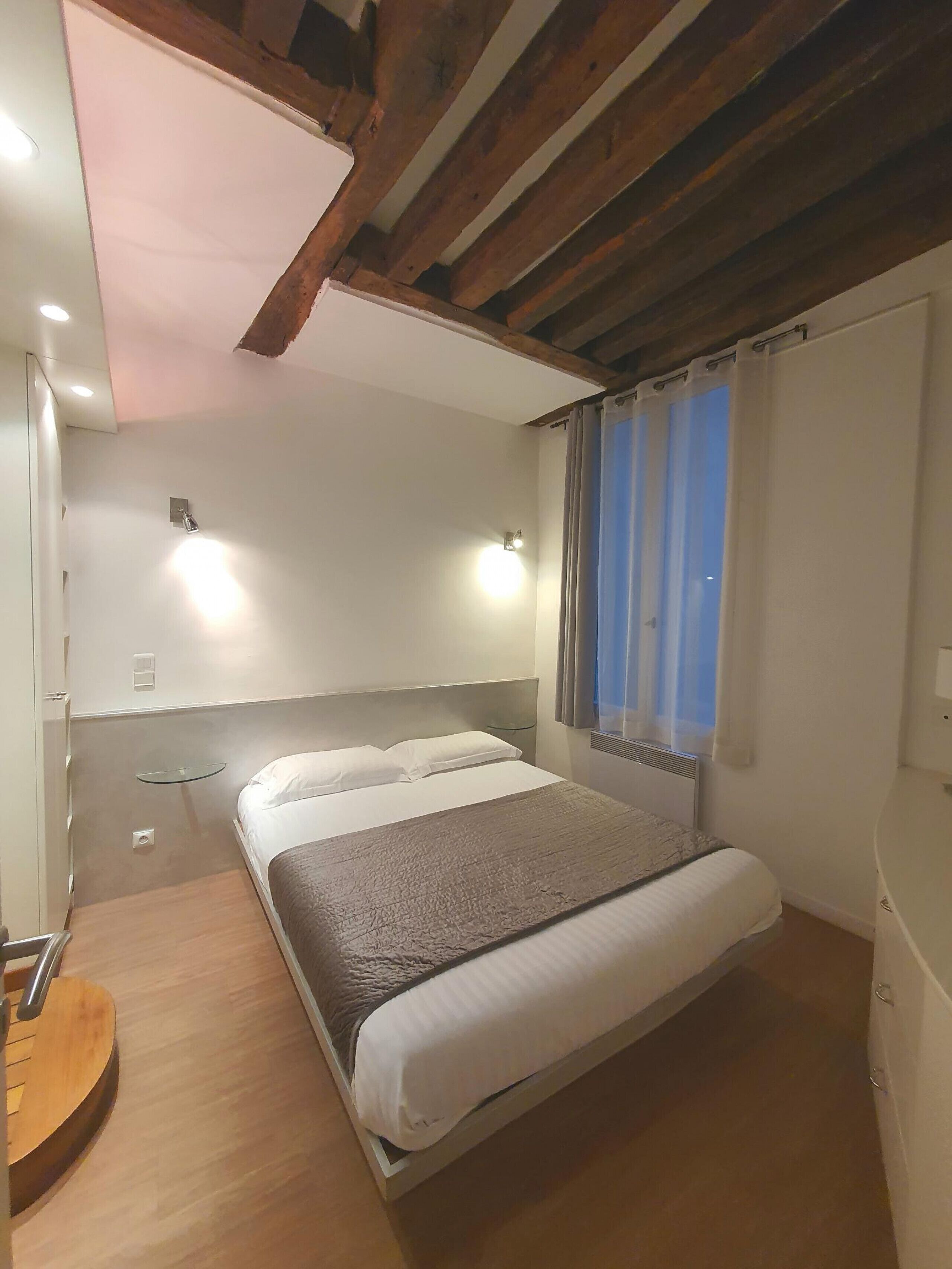 A bedroom with a bed, a bedside table, a small shelf, and a window with curtains.
