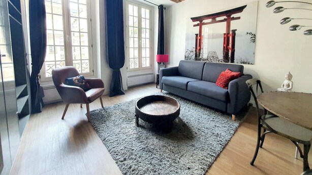 Un salon comprenant un canapé, une petite table ronde, une chaise, un tapis et une fenêtre avec des rideaux.