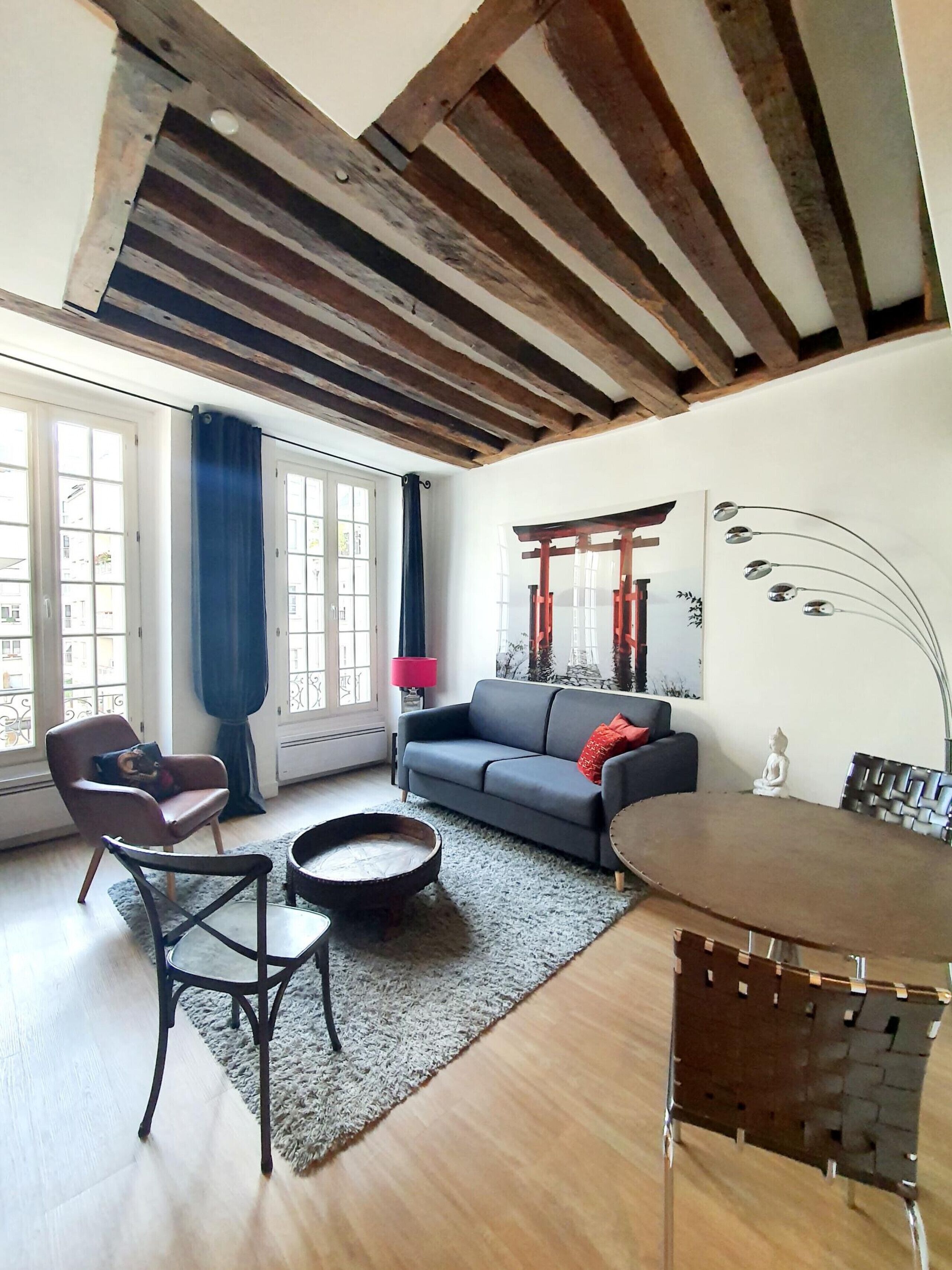 A living room with a wooden ceiling, a grey sofa, a round coffee table, a small dining table, and a chair.