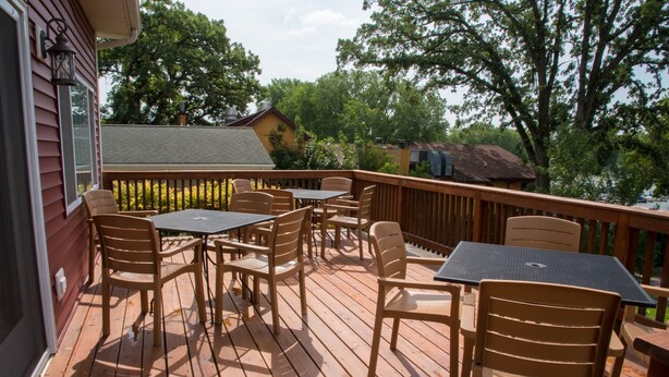 Una terraza de madera con mobiliario exterior, incluyendo sillas y mesas, con vista a un cuerpo de agua rodeado de árboles.