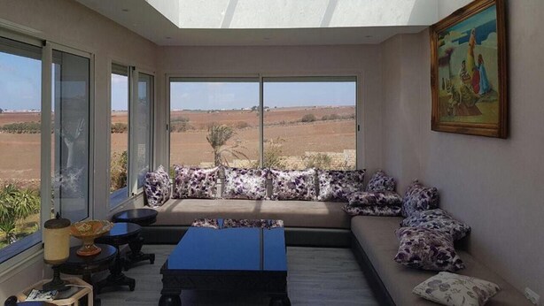 A spacious room with a large window offering a view of the countryside. There is a sofa with patterned cushions and a blue coffee table.