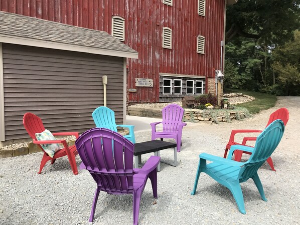 Un patio de grava con sillas Adirondack de colores y una mesa frente a un granero rojo con un letrero que indica un restaurante.