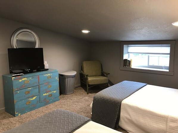 Un dormitorio con cama, una silla, cómoda, televisor, ventana y cubo de basura.
