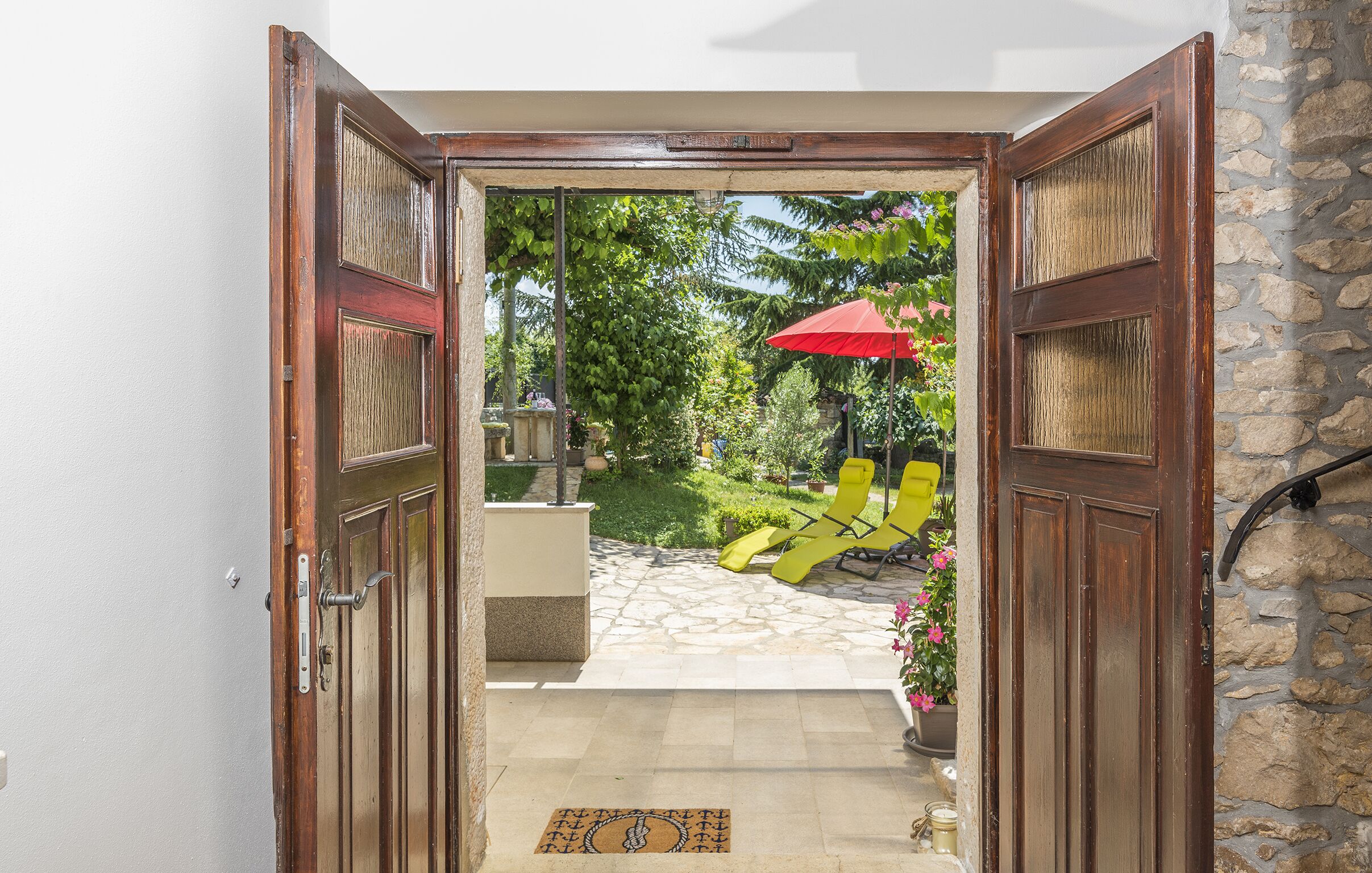 Une porte en bois ouverte donne sur un patio dallé de pierres, où se trouvent des fauteuils de jardin jaunes et un parasol rouge.