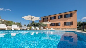 Une piscine avec des chaises longues et des parasols, un bâtiment à deux étages doté de balcons, et un ciel d’un bleu limpide.