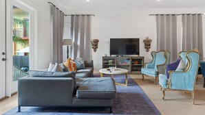 Una sala de estar con un sofá gris, una mesa de centro y dos sillones azules. Un televisor está montado en la pared, y hay un mueble bajo él.