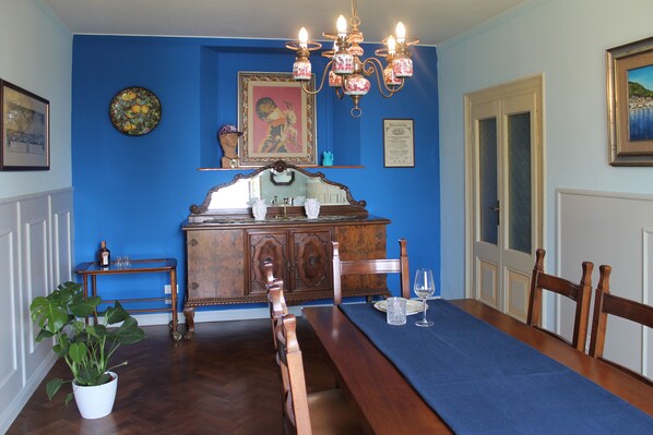 Comedor con mesa de madera dispuesta para cuatro personas, lámpara de araña y pared azul decorada con cuadros.