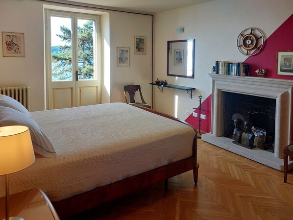 A bedroom with a bed, a chair, a fireplace, and a window with a view of trees.