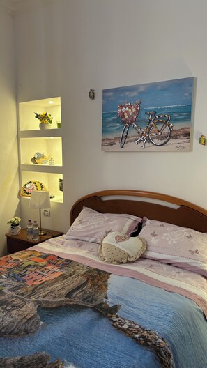 A bedroom with a bed, a nightstand, a lamp, a wall-mounted shelf with decorative items, and a painting of a bicycle on a beach.