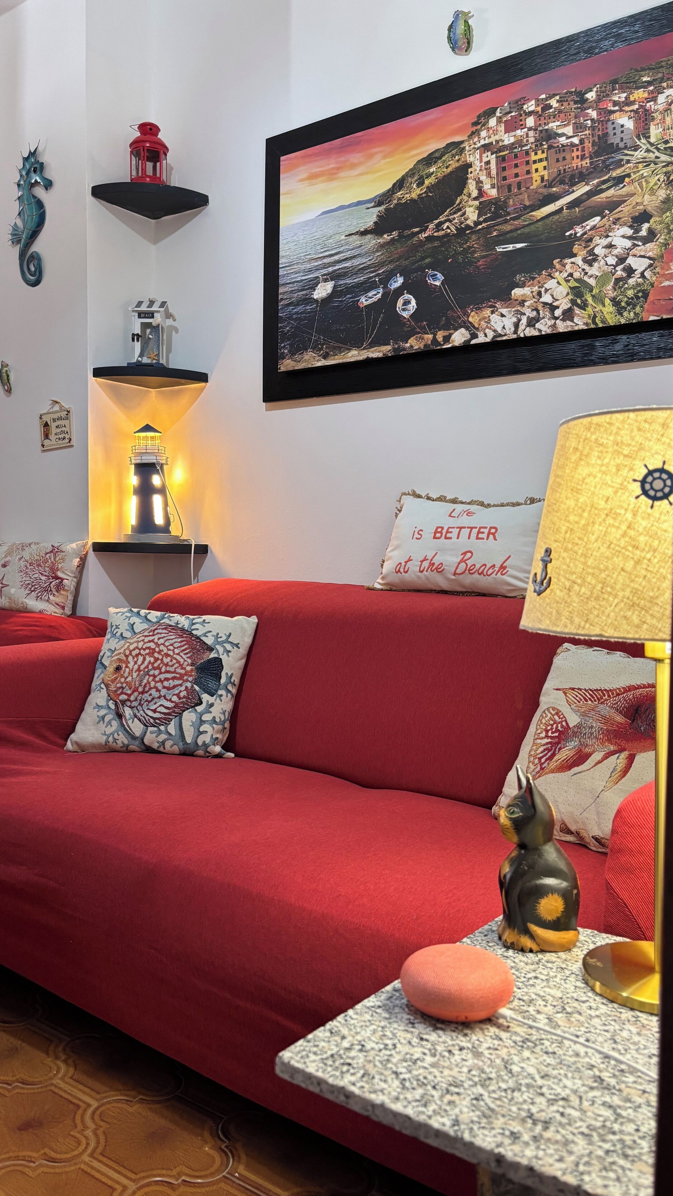 Ein rotes Sofa mit dekorativen Kissen, ein kleiner Tisch mit einer Lampe, ein Wandregal mit einer Laterne und ein gerahmtes Bild einer Küstenstadt.