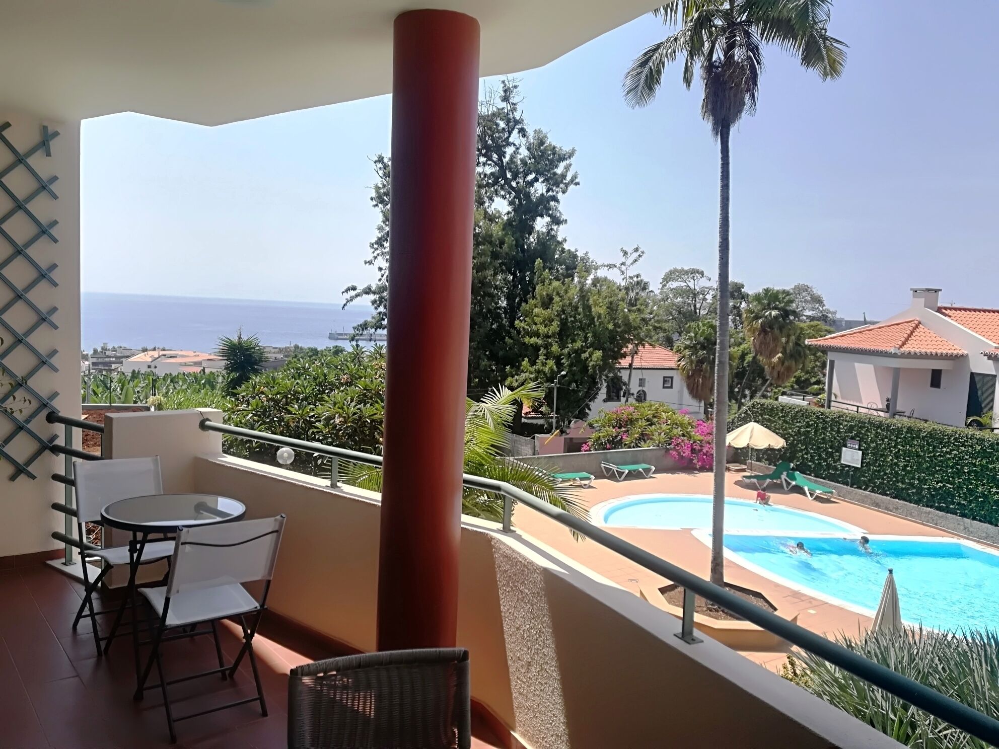 A balcony with a table and chairs, a view of a swimming pool, and a palm tree.
