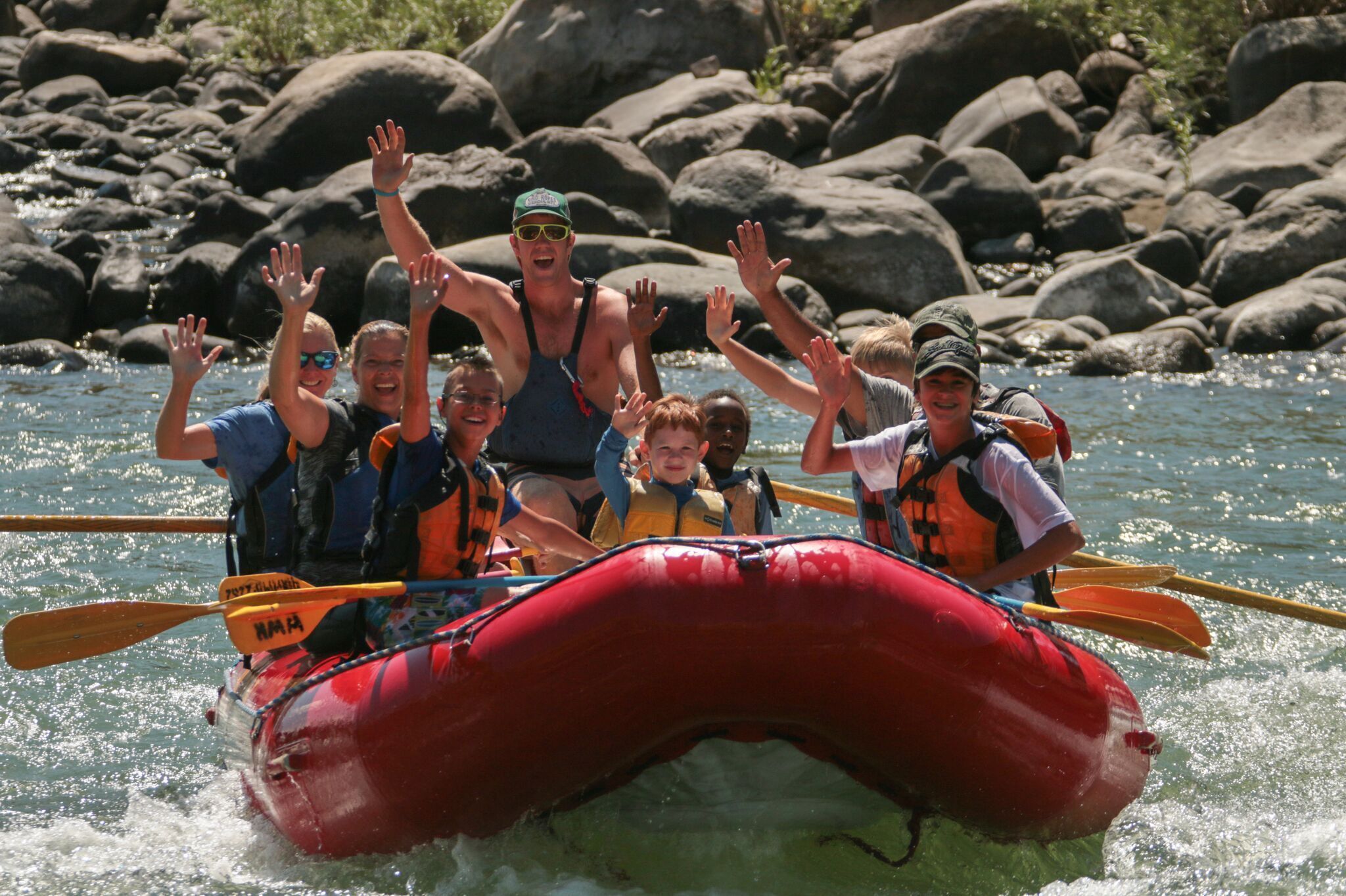 Een groep mensen in een rode vlot met peddels, met reddingsvesten, op een rivier omringd door rotsen.