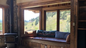 Ein gemütliches Blockhaus-Interieur mit Kamin aus Stein, einer Bank mit Kissen und Blick auf einen bewaldeten Hang.