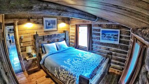 Ein Schlafzimmer in einer Blockhütte mit einem Bett, zwei Nachttischen, einem Fenster und einem an der Wand hängenden Bild.